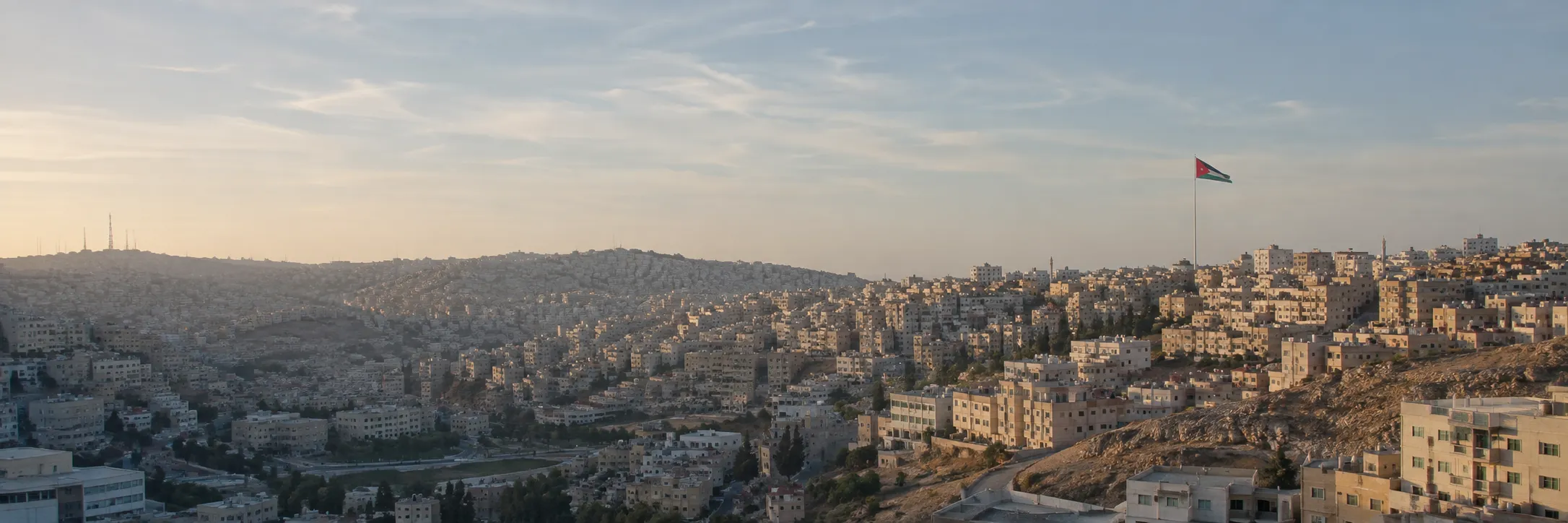 منظر بانورامي لمدينة عمّان عند شروق الشمس، ويظهر العلم الأردني فوق أبنية المدينة الحجرية.